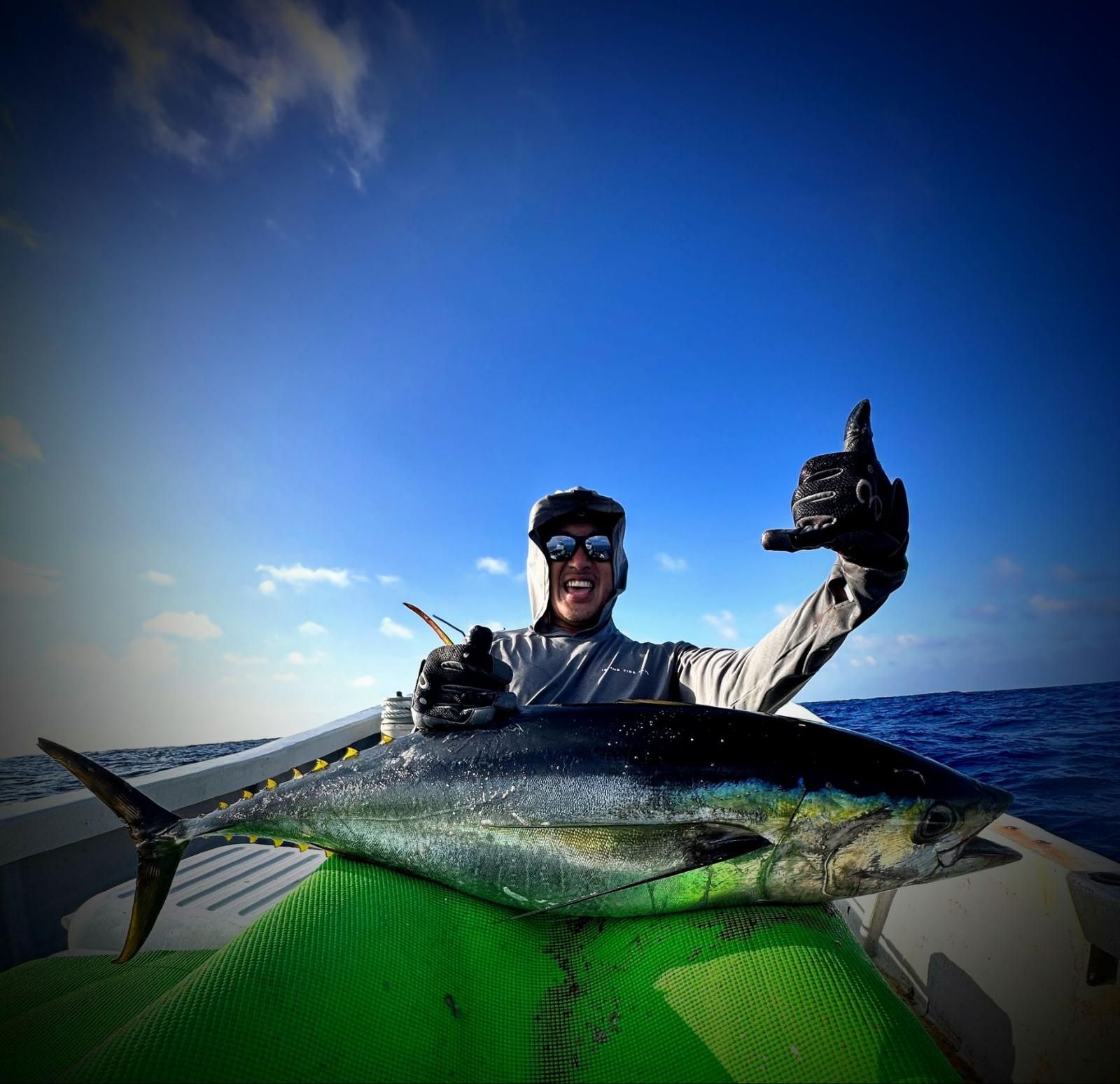 Angler grinning on a small boat beside a freshly landed yellowfin tuna under a deep blue sky